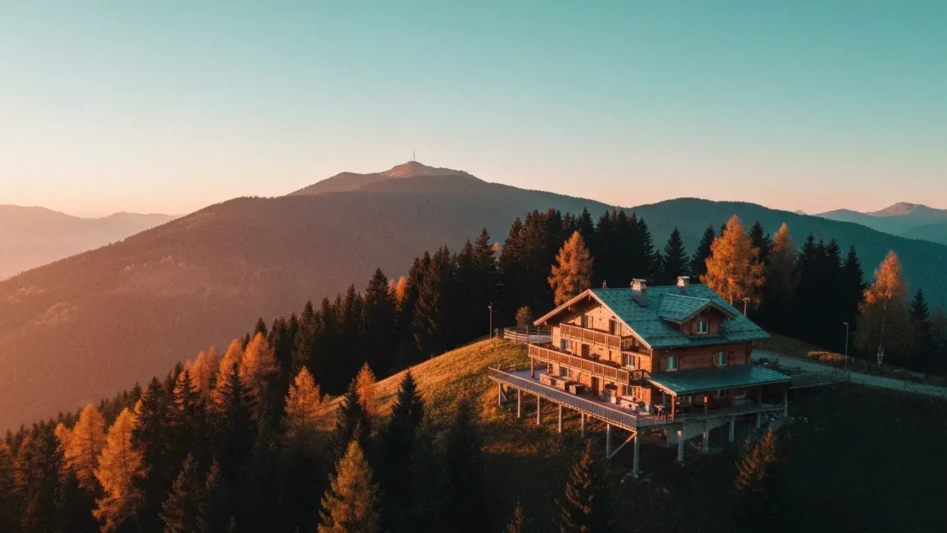 An sweeping aerial photograph of a mountain chalet perched above the treeline, at golden hour