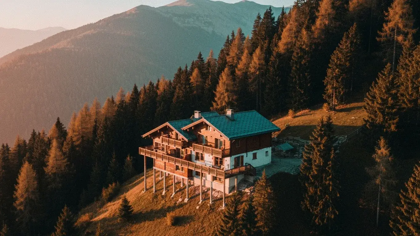 An aerial drone photograph of a mountain chalet perched above the treeline, in soft morning light