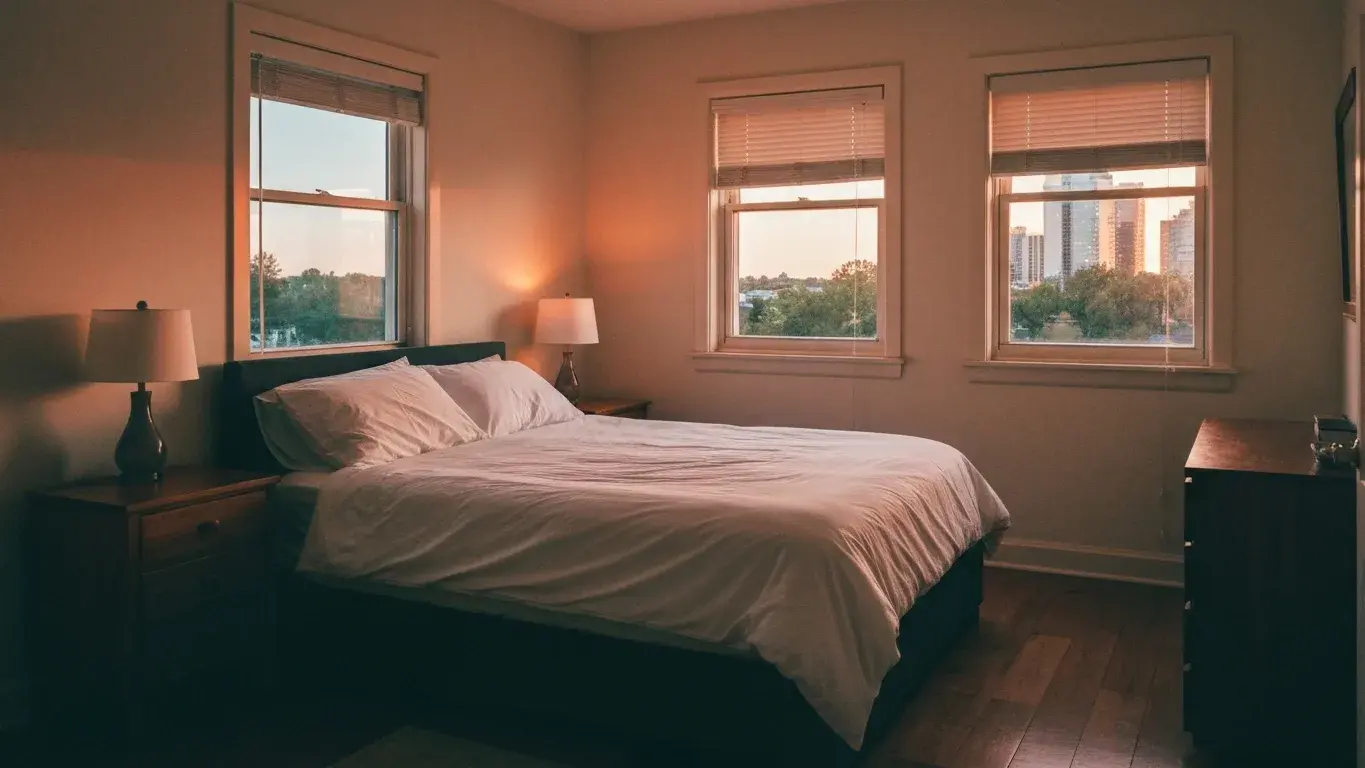 wide interior photograph of a sunlit primary bedroom with a made bed with crisp white linens, lit by table lamps at dusk