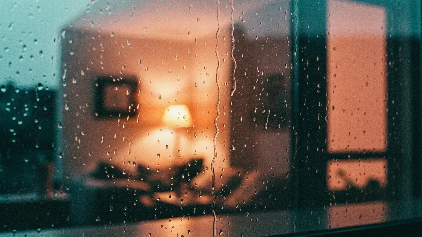 evocative photograph of rain streaking a window with a cozy lit interior beyond, peaceful and quiet